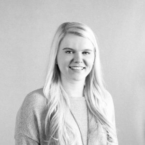 A black and white portrait of a young woman with long blonde hair, smiling. She is wearing a light-colored sweater and a necklace, against a plain background.